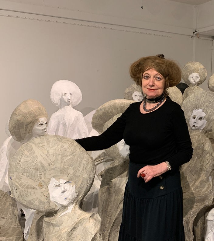 Marianne Pitzen in einem schwarzem Kleid. Sie steht vor grauen Skulpturen, die ihr ähnlich sehen.