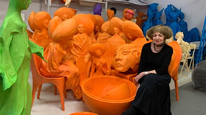 Marianne Pitzen mit schwarzem Kleid, sitzt vor einigen orangenen Skulpturen.