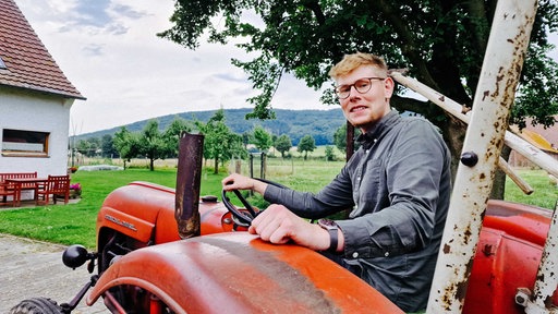 Mathis Lemke sitzt lächelnd auf einem alten Traktor, der auf seinem Hof und schaut über die Schulter in die Kamera. Im Hintergrund sind Landschaft und Berge.