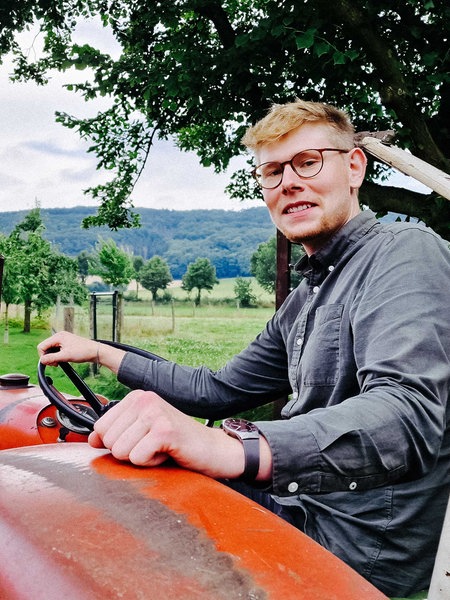 Mathis Lemke sitzt lächelnd auf einem alten Traktor, der auf seinem Hof und schaut über die Schulter in die Kamera. Im Hintergrund sind Landschaft und Berge.