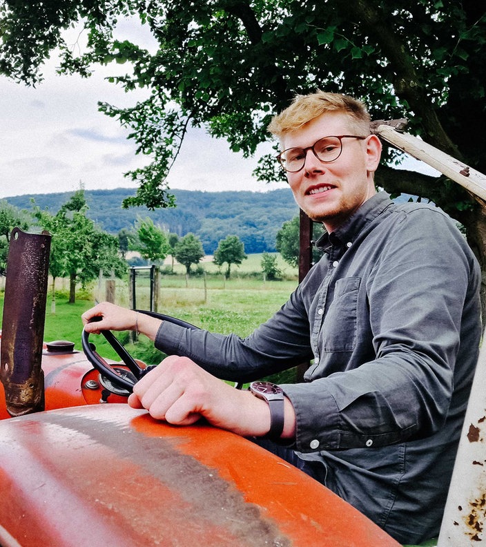 Mathis Lemke sitzt lächelnd auf einem alten Traktor, der auf seinem Hof und schaut über die Schulter in die Kamera. Im Hintergrund sind Landschaft und Berge.