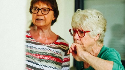 Eine ältere Frau mit weißen Haaren und roter Brille schaut nachdenklich. Eine zweite Frau mit bunten Oberteil neben ihr