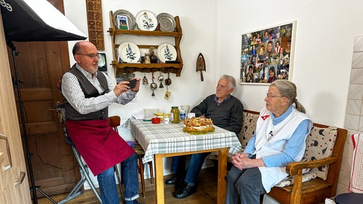 Oma Agnes sitz am Küchentisch, vor ihr steht ein Kuchen. Sie wird gefilmt.