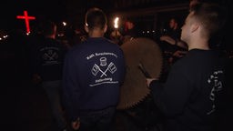 Teilnehmer bei der Krachnacht in Hallenberg schlagen auf ein Sägeblatt, außerdem sind Fackeln und ein knallrot leuchtendes Kreuz im Zug zu sehen.