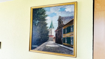 Ein Ölgemälde hängt an einer Wand. Es zeigt eine Kirche im Zentrum, sowie ein Wohnhaus mit grünen Fenstern am rechten Bildrand