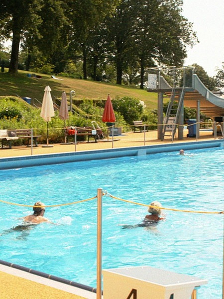 Das Schwimmbecken eines Freibads mit Liegen am Beckenrand, einer kleinen Rutsche im Hintergrund sowie einigen Schwimmern im Wasser.