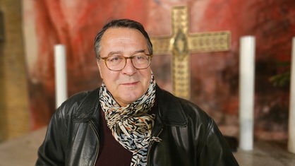 Pfarrer Hans Mörtter steht vor einem Kreuz und lächelt leicht in die Kamera
