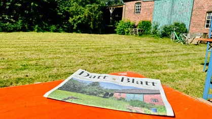 Zu sehen ist eine Zeitung, die auf einer Decke auf einer Wiese liegt.