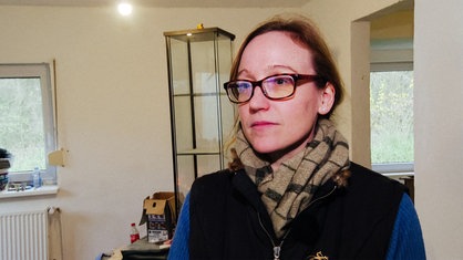 Eine Frau mit Brille und Schal steht in ihrem zu renovierenden Wohnzimmer