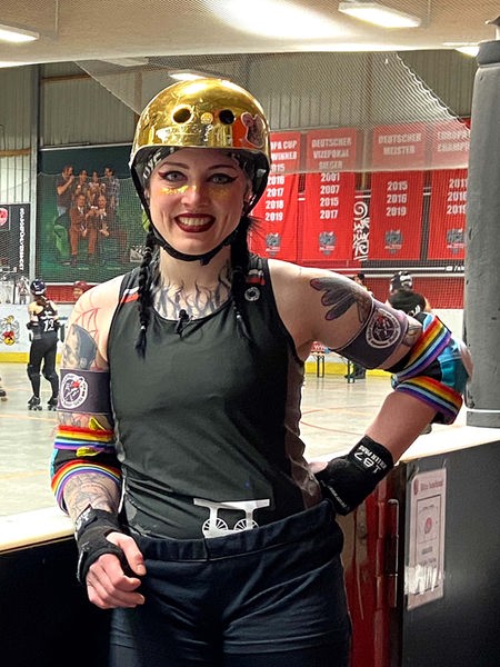 Melina Brinkmann steht an der Bande einer Hockeyarena, hinter ihr spielen Frauen Roller Derby