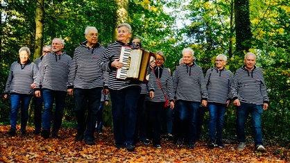 Der Shanty-Chor steht im Wald und singt gemeinsam. Sie tragen alle blau-weiß gestreifte Oberteile, vorne steht eine Frau und spielt Akkordeon.