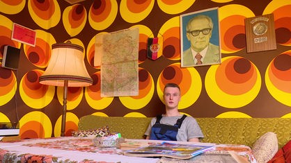 Dennis Vette in seinem DDR-Wohnzimmer: An den Wänden eine 70er-Jahre-Tapete mit braun-organgenen Kreisen und ein Porträt von Erich Honecker