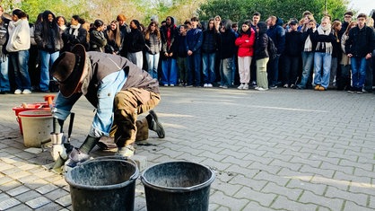 Ein Mann kniet am Boden und verlegt Stolpersteine, im Hintergrund sehen viele Schüler zu