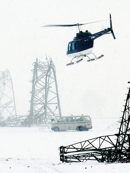 Umgeknickte Strommasten auf einem eingeschneiten Feld. Darüber fliegt ein Hubschrauber. Im Hintergrund sind Fahrzeuge zu sehen.