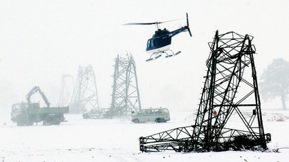 Umgeknickte Strommasten auf einem eingeschneiten Feld. Darüber fliegt ein Hubschrauber. Im Hintergrund sind Fahrzeuge zu sehen.