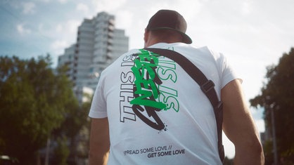 Eine Person steht vor einem Hochhaus und trägt ein T-Shirt, auf dem "This is Gelsen" steht.