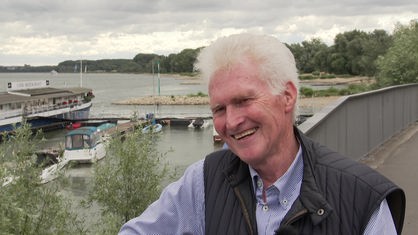 Grauhaariger Mann lacht in Kamera, Rheinufer im Hintergrund