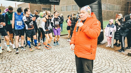 Peter Berghaus mit einem Mikro in der Hand steht vor der Startlinie des Rennens. Im Hintergrund stehen bereits einige Läufer und Läuferinnen, die auf sein Signal warten