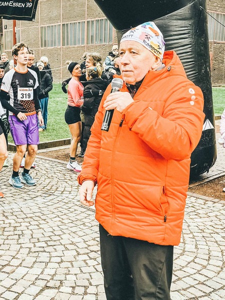 Peter Berghaus mit einem Mikro in der Hand steht vor der Startlinie des Rennens. Im Hintergrund stehen bereits einige Läufer und Läuferinnen, die auf sein Signal warten