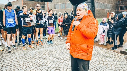 Links eine Gruppe von Läufern. Rechts Peter Berghaus in organgener Jacke, der ein Mikrofon in der Hand hält.