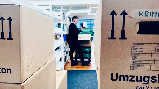 Eine Frau in dunkler Kleidung und mit kurzen Haaren zwischen Umzugskartons. Sie packt Bücher ein.