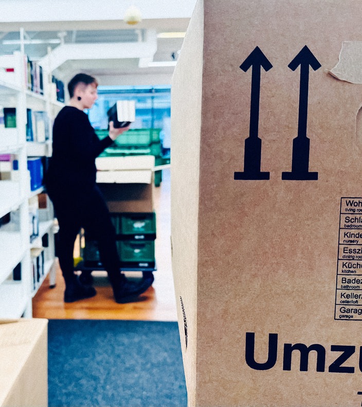 Eine Frau in dunkler Kleidung und mit kurzen Haaren zwischen Umzugskartons. Sie packt Bücher ein.