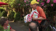 Mann mit gelber Kappe bietet einem sitzenden Mann Zeitschriften zum Verkauf an