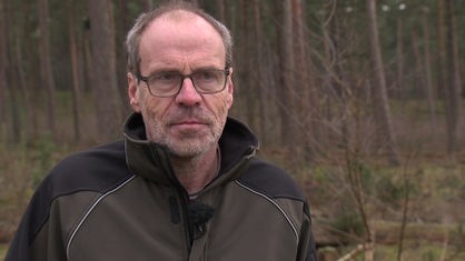 Mann mit Brille und Arbeitsjacke steht in einem Waldgebiet und spricht in Kamera