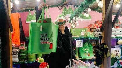 Christoph Scheiding steht hinter seinem Weihnachtsmuffel-Stand. Er hält eine Weihnachtsmuffel-Tasche hoch und trägt eine dazu passende Brille.