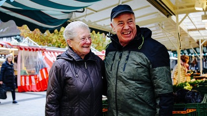 Stammkundin Frau Scholz und Volker Schling lächelnd Arm in Arm auf dem Markt.