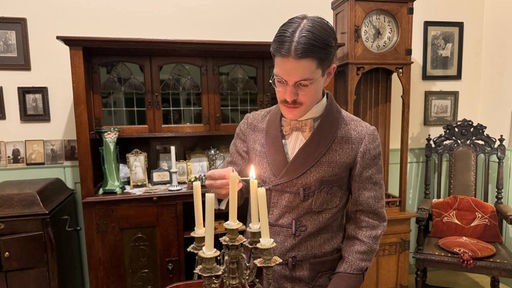 Ein Mann steht in einem Wohnzimmer, welches mit alten Möbeln eingerichtet ist und zündet eine Kerze an.