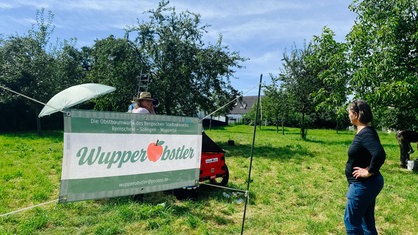 Ein Banner auf einer grünen Wiese, auf dem "Wupperobstler" steht. Davor und dahinter stehen zwei Frauen