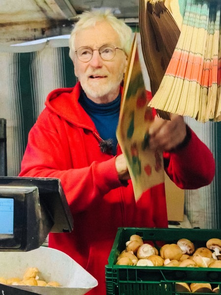 Ein Mann in einer roten Jacke an einem Gemüsestand, er ist dabei, Champignons abzuwiegen