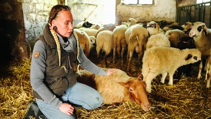 Vicky Diemel sitzt auf dem Boden ihres Schafstalles. Mit der linken Hand streichelt sie ein Schaf, dass neben ihr liegt.