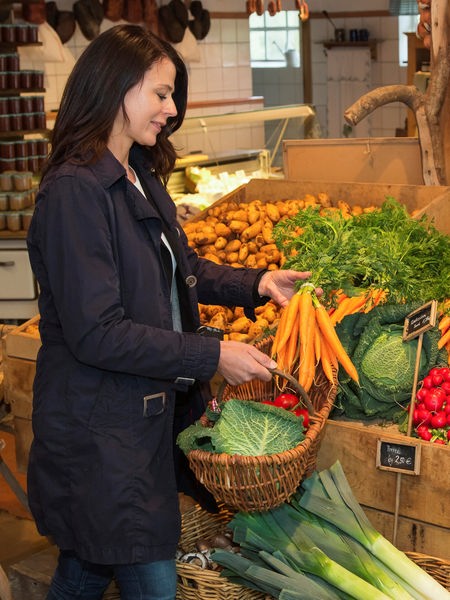 Eine Frau steht in einem Hofladen und verstaut vor einem Gemüseregal Einkäufe in ihren Korb