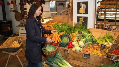 Eine Frau steht in einem Hofladen und verstaut vor einem Gemüseregal Einkäufe in ihren Korb