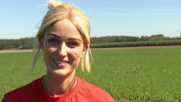 Junge Frau mit blonden Haaren steht lächelt in Kamera, sie trägt ein rotes T-Shirt und steht auf einem Feld