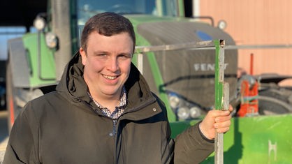 Ein Landwirt mit braunen Haaren steht an einem grünen Trecker. 