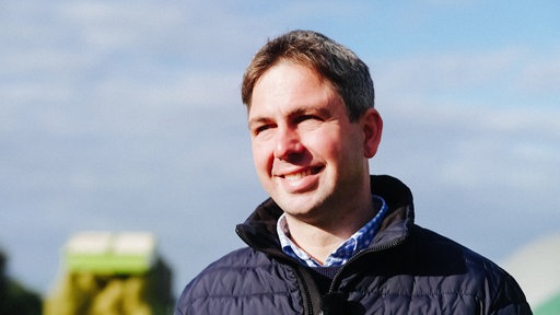 Ein Mann mit kurzen Haaren und blauer Jacke lächelt in die Kamera.