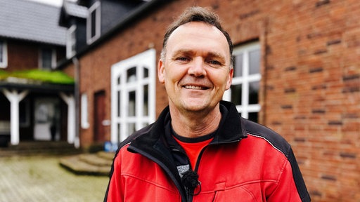 Ein Mann mit roter Regenjacke, der vor einem geklinkerten Haus steht.