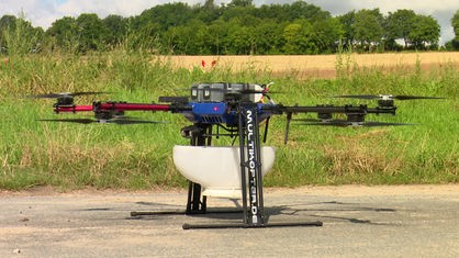Eine große Drohne, an der ein weißer Behälter befestigt ist, steht vor einem Feld.