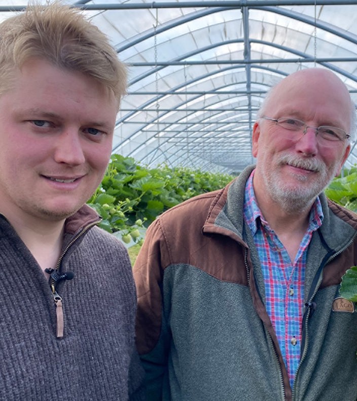 Zu sehen sind zwei Herren, die zwischen Erdbeerpflanzen stehen und in die Kamera lächeln.
