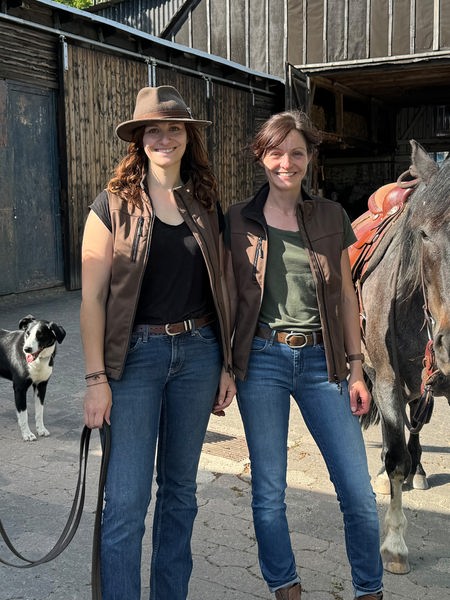 Zwei Frauen stehen zwischen zwei Pferden vor einem Stall und schauen in die Kamera.