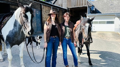 Zwei Frauen stehen zwischen zwei Pferden vor einem Stall und schauen in die Kamera.