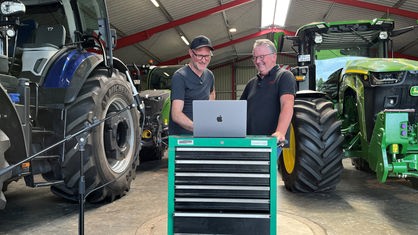 Musikproduzent Henning Verlage auf der linken Seite und Hubert Wilmer auf der rechten, stehen in einer Wagenhalle mit mehreren Traktoren im Hintergrund. Vor ihnen steht ein Werkzeugwagen, auf dem ein Laptop steht.