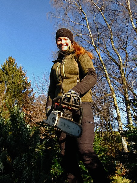 Sarah Neßhöver aus der Froschperspektive in Arbeitskleidung und mit Kettensäge auf der Weihnachtsbaumplantage