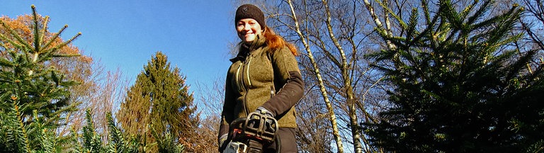 Sarah Neßhöver aus der Froschperspektive in Arbeitskleidung und mit Kettensäge auf der Weihnachtsbaumplantage