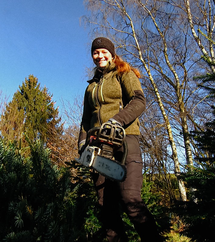 Sarah Neßhöver aus der Froschperspektive in Arbeitskleidung und mit Kettensäge auf der Weihnachtsbaumplantage