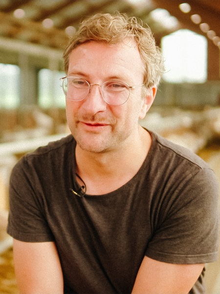 Ein junger Mann mit bräunlichen Haaren und einer Brille. Im Hintergrund ein Stall mit vielen Ziegen