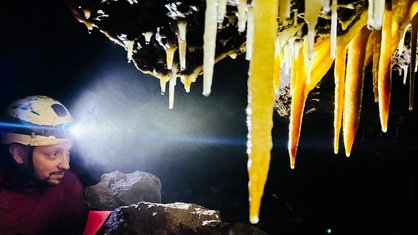 Ein Mann mit Helm und Stirnlampe kriecht in einer Höhle, im Vordergrund sind große Stalagtiten